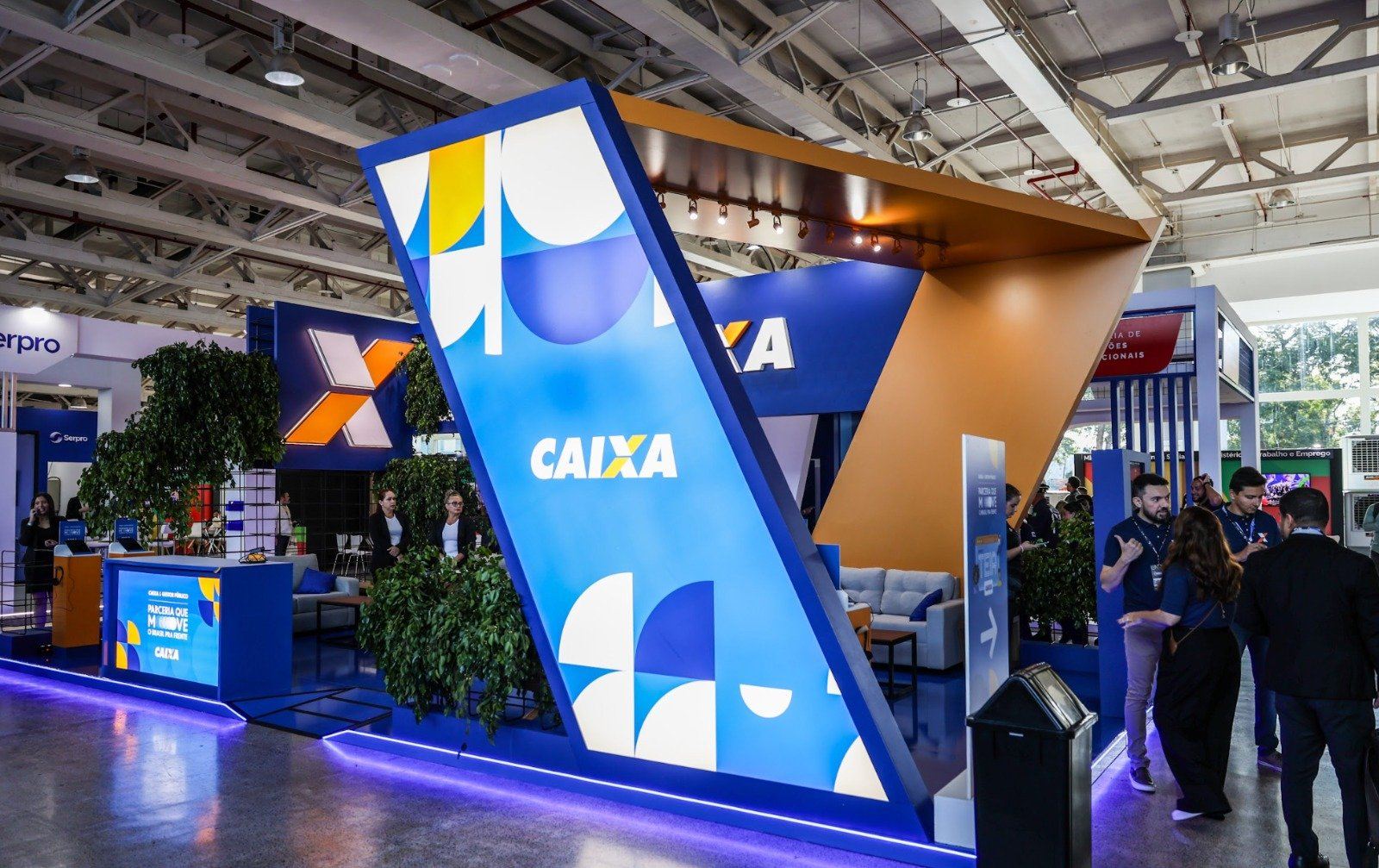 Built Caixa booth: a tilted blue A-frame with an orange canopy, visitors talking in front